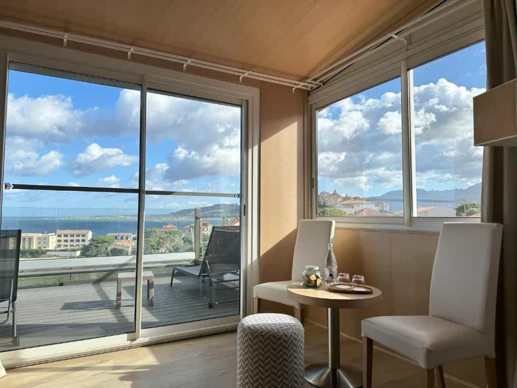 Hotel room interior with chairs and balcony view of coastal town and sea under blue sky