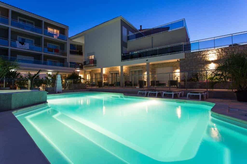 Illuminated swimming pool at Hotel Revellata & Spa at dusk with modern hotel buildings.