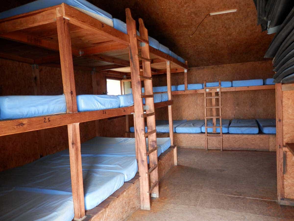 Wooden bunk beds with blue mattresses inside a simple shelter with particle board walls and ceiling.