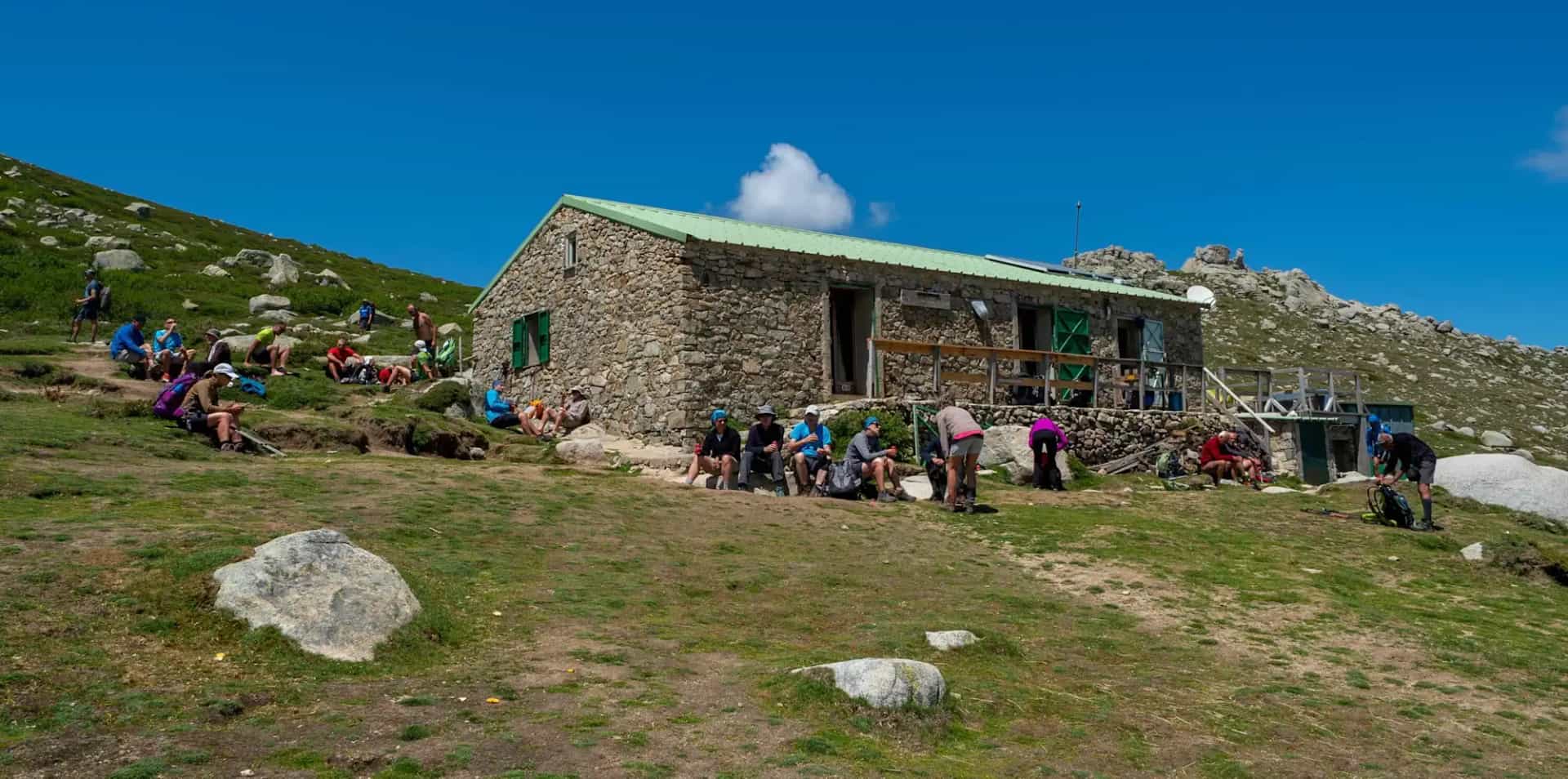 Hikers resting outside a stone mountain refuge with a green roof under a bright blue sky.