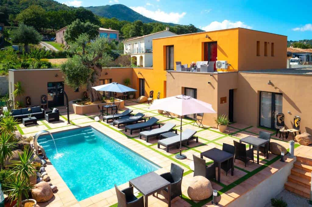 Swimming pool patio with lounge chairs, umbrellas, and modern buildings against green hills.