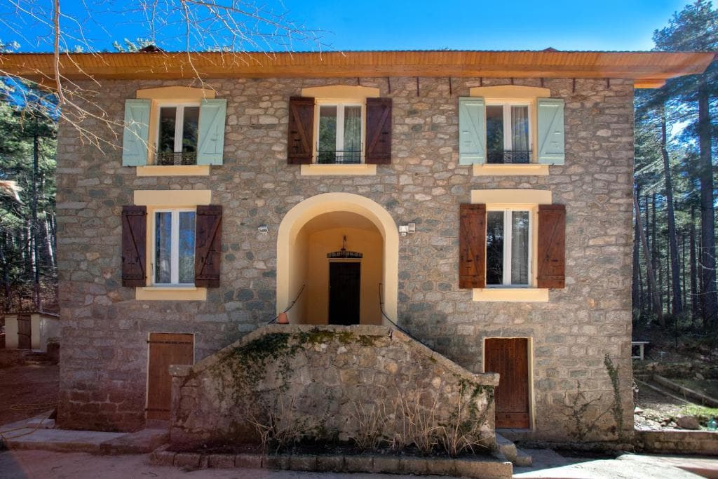 Stone house with arched entrance and wooden shutters next to a dense pine forest, Casa Alta.