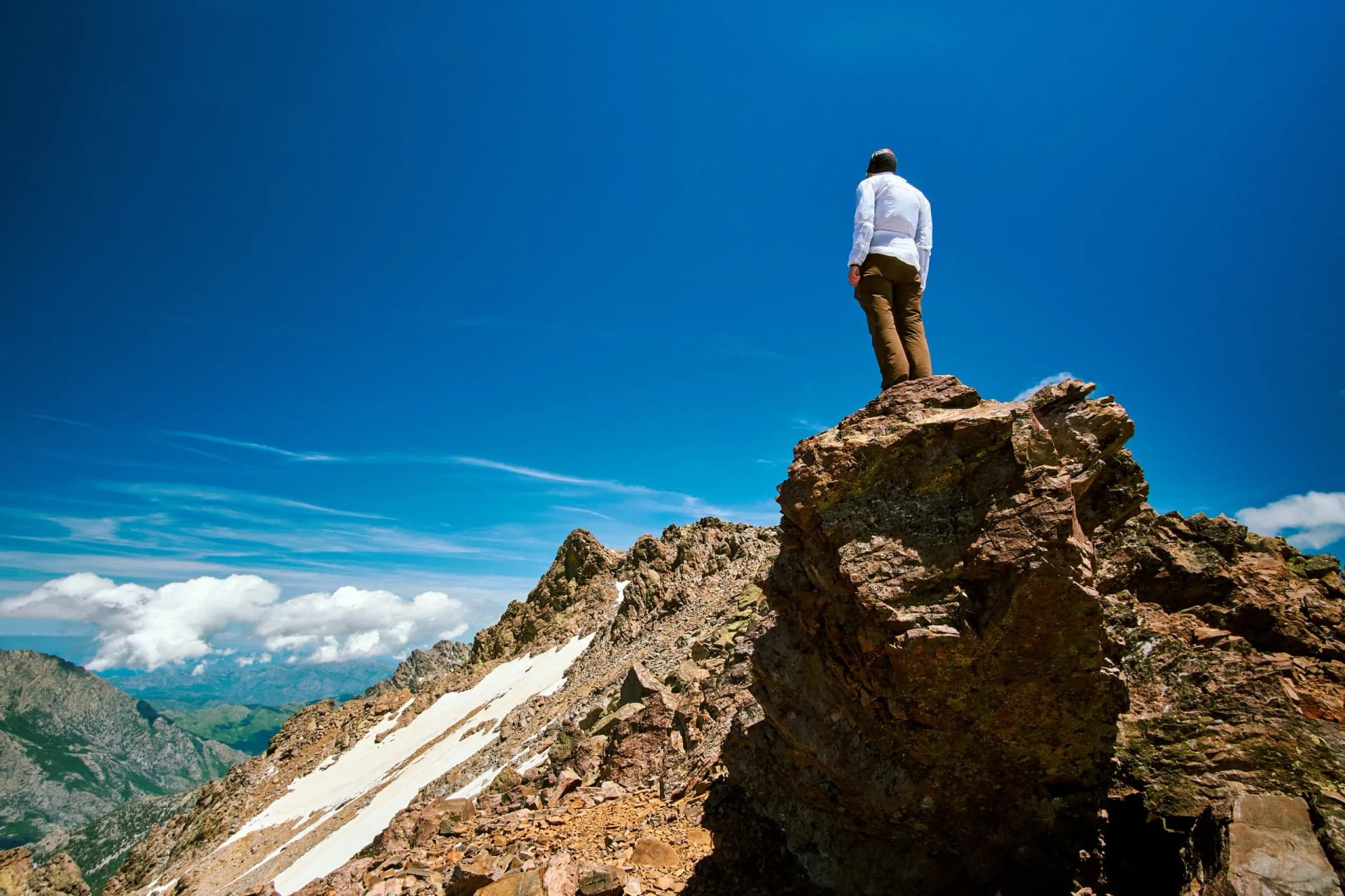Hiker standing on rocky summit overlooking mountains with patches of snow under bright blue sky.
