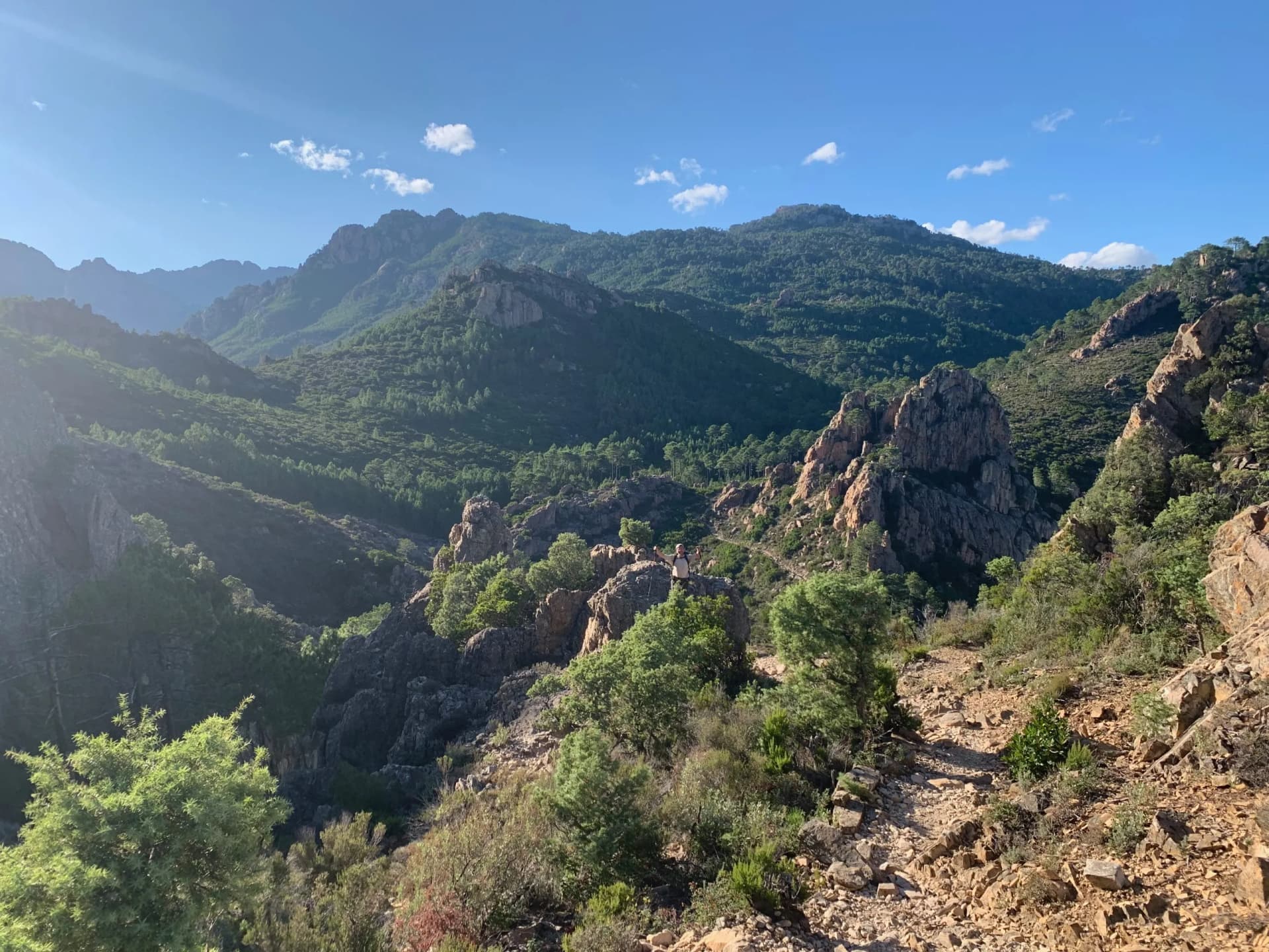 Hiking on rocky trail with scrub brush, mountains, and blue sky in Conca, Corsica.