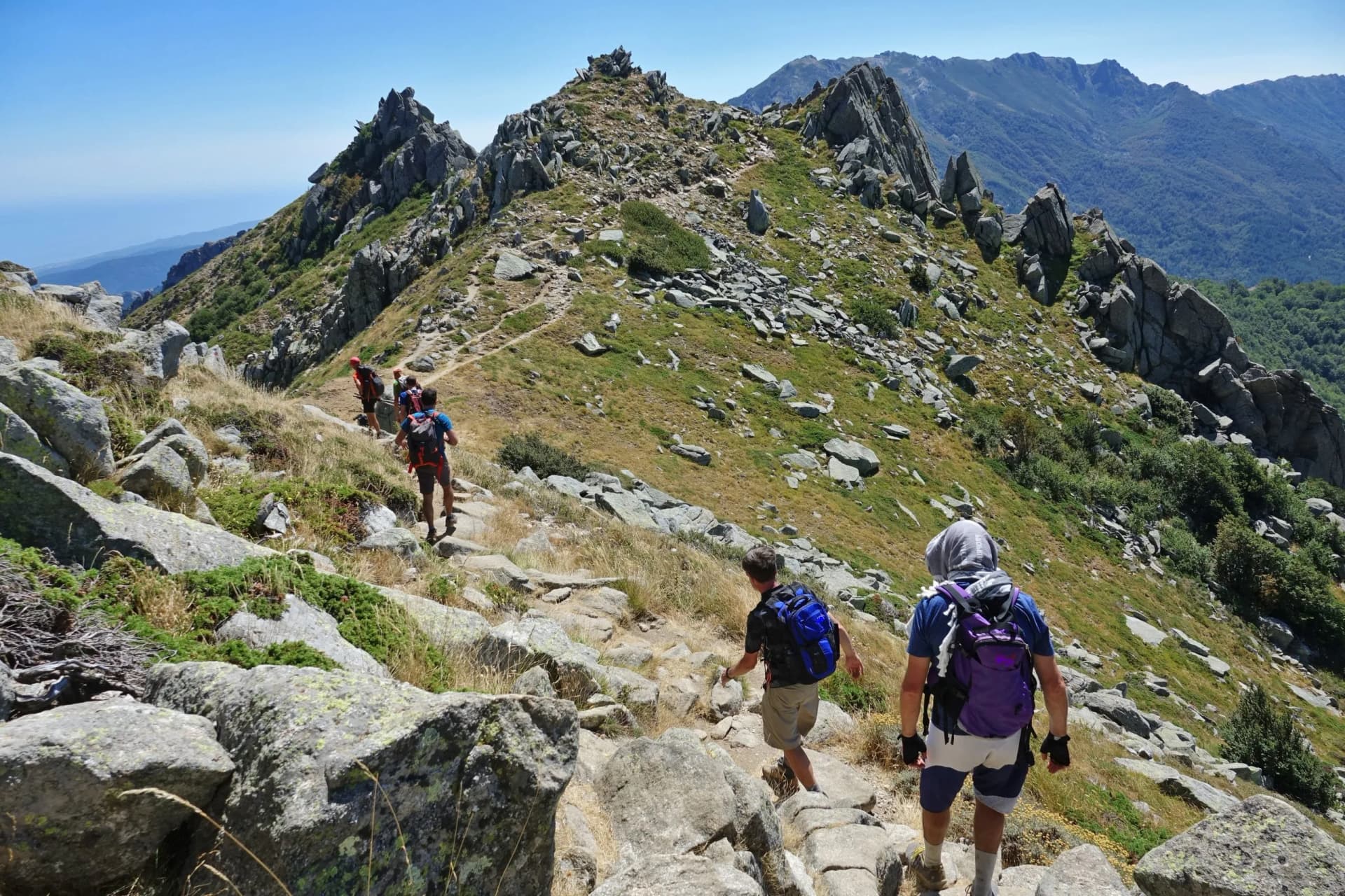 Hikers ascending a rocky mountain trail toward sharp peaks, with the sea visible in the distance, Corsica.