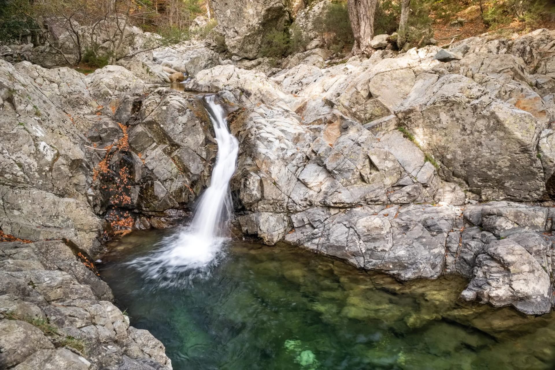 Waterfall cascades over rocks into a green pool in the forest of Vizzavona along the GR20 trail in Corsica.